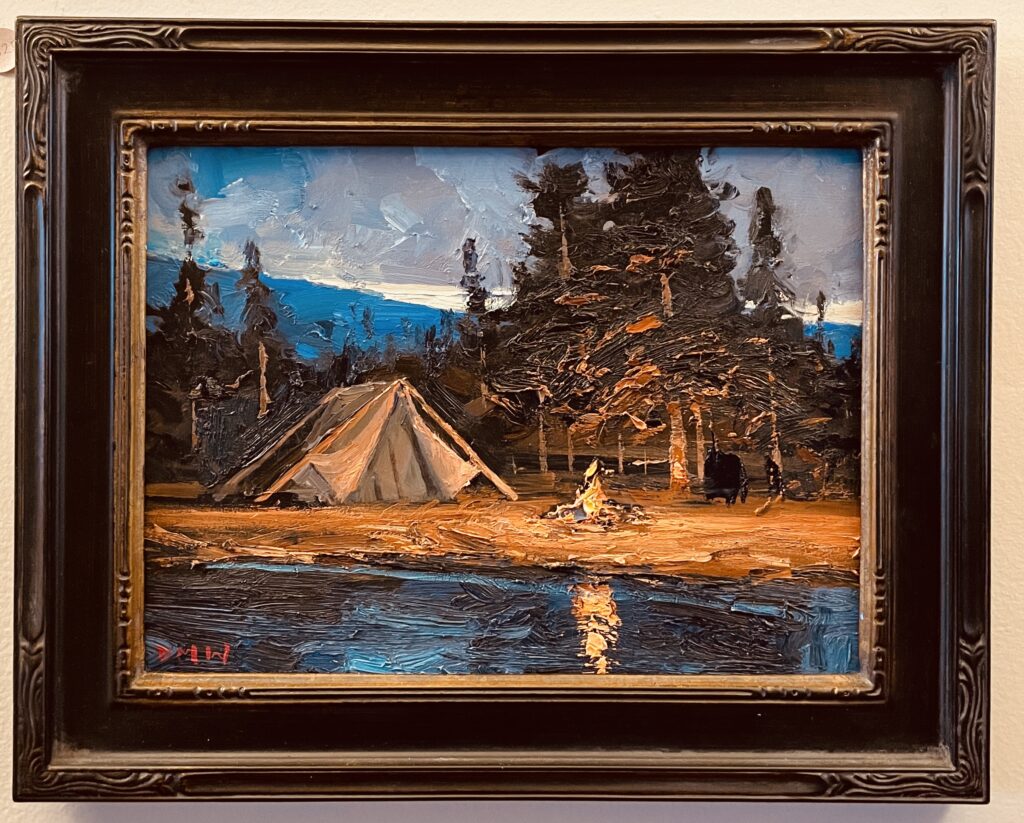 In an ADK oil painting with built-up texture, there is the refection of a blazing campfire in the foreground water. A canvas tent rests on shore next to the fire. There are lit-up pines and a blue sky with clouds in the background.