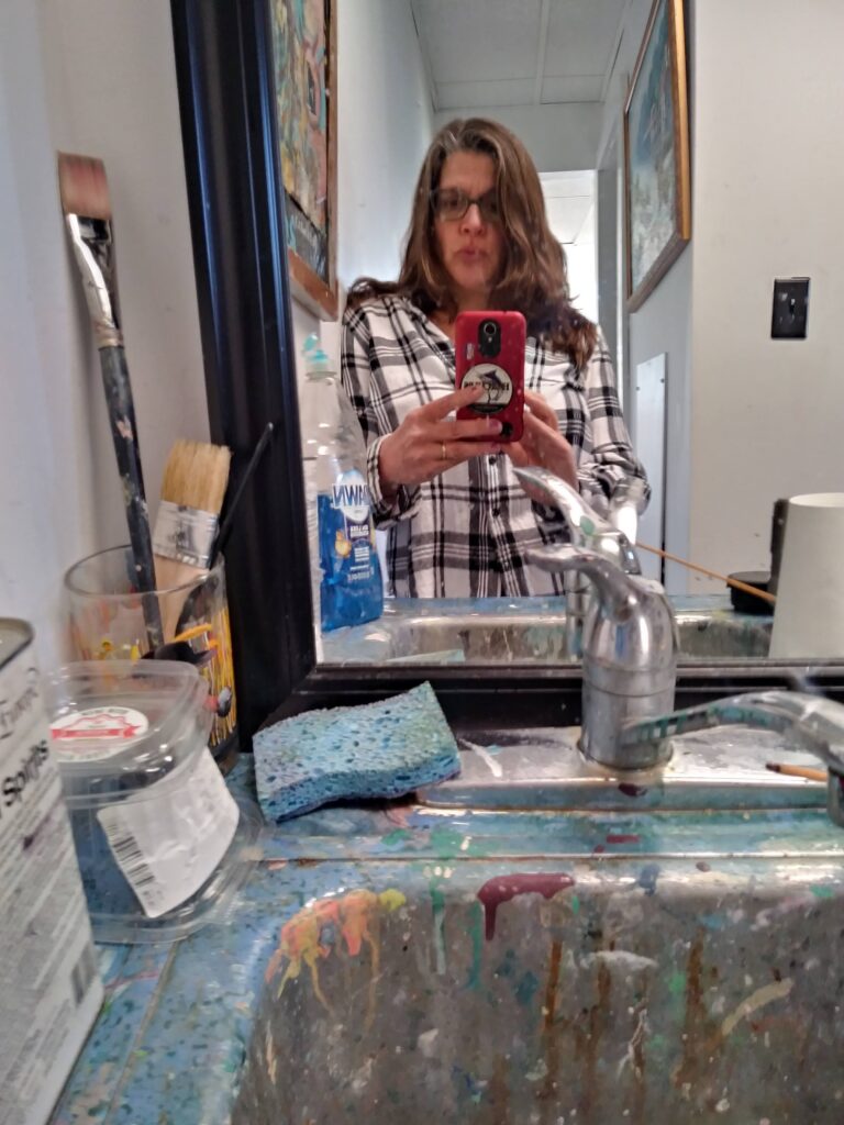 Self-Portrait in Mirror. Woman stands in front of a mirror showing her reflection. A sink appears in the foreground with paint splatters, brushes other painting tools.